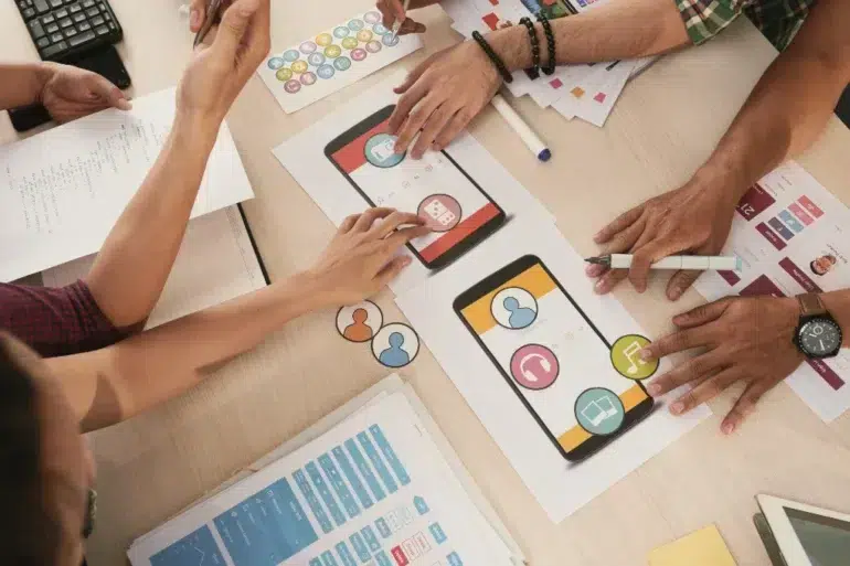 Overhead view of diverse hands collaborating on smartphone app designs at a desk. Colorful icons and sketches suggest teamwork and creativity.