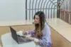 A young woman focusing on work with a laptop and notepad in a modern indoor setting.