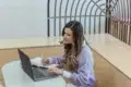 A young woman focusing on work with a laptop and notepad in a modern indoor setting.