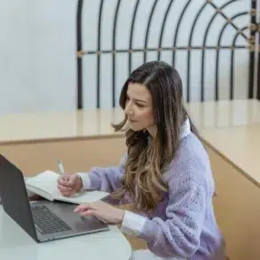 A young woman focusing on work with a laptop and notepad in a modern indoor setting.