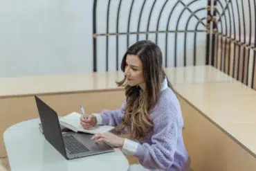 A young woman focusing on work with a laptop and notepad in a modern indoor setting.