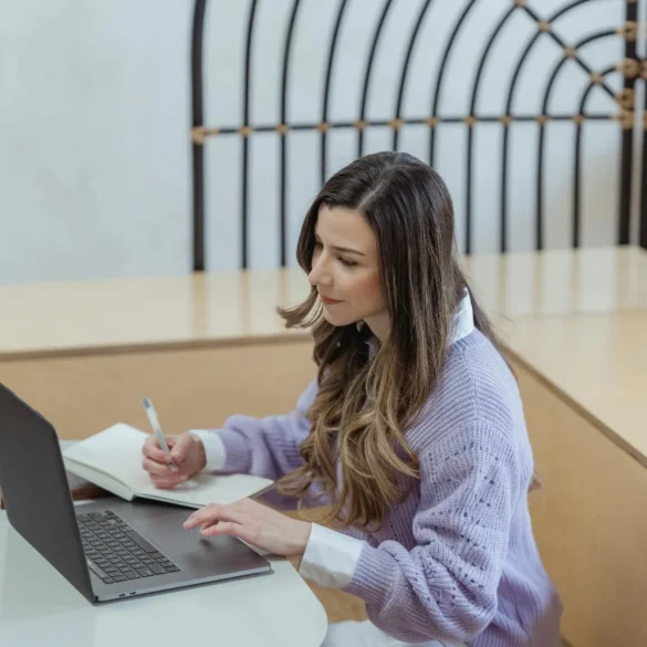 A young woman focusing on work with a laptop and notepad in a modern indoor setting.