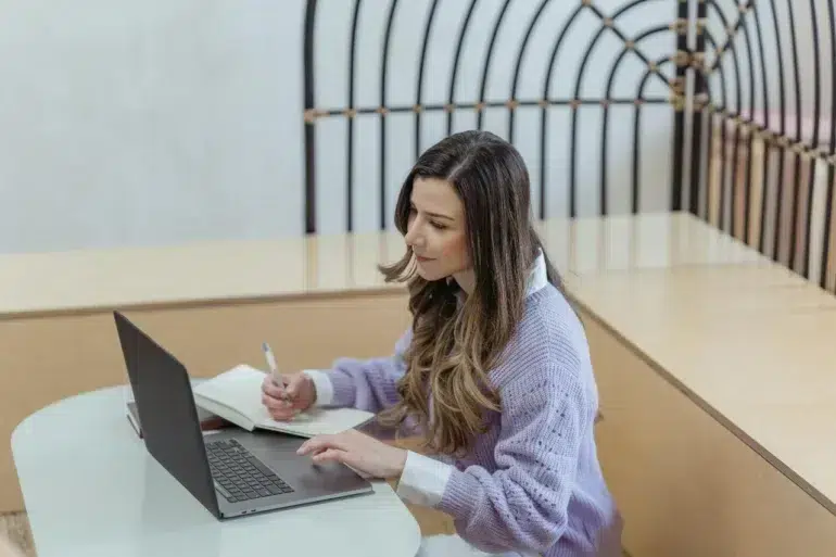 A young woman focusing on work with a laptop and notepad in a modern indoor setting.