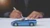 A small blue convertible toy car is in focus on a table. In the blurred background, a person in business attire signs a document, suggesting an auto-related context.