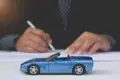 A small blue convertible toy car is in focus on a table. In the blurred background, a person in business attire signs a document, suggesting an auto-related context.