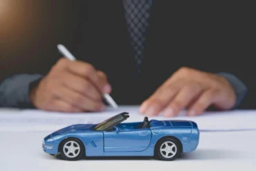 A small blue convertible toy car is in focus on a table. In the blurred background, a person in business attire signs a document, suggesting an auto-related context.