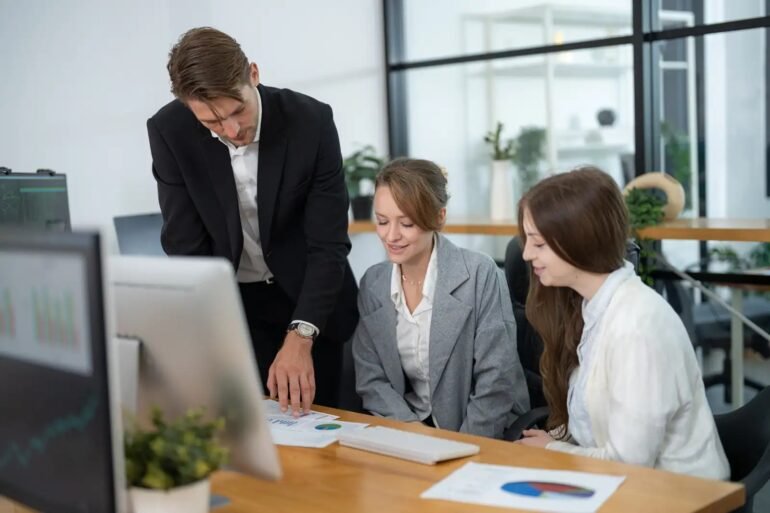 Three business professionals reviewing charts and data at a desk in a modern office setting.