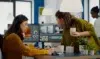 Two women collaborating in an office, focused on video editing software on a dual monitor setup. One holds a coffee, creating a productive atmosphere.