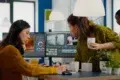 Two women collaborating in an office, focused on video editing software on a dual monitor setup. One holds a coffee, creating a productive atmosphere.