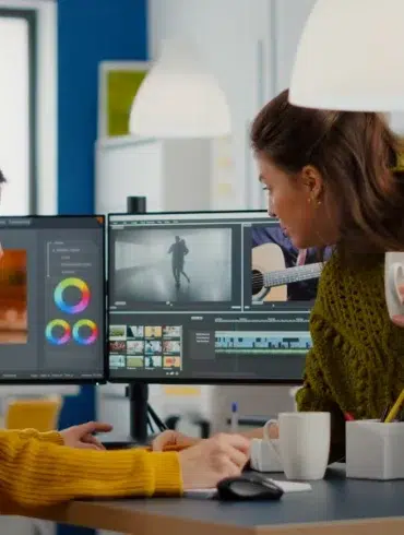 Two women collaborating in an office, focused on video editing software on a dual monitor setup. One holds a coffee, creating a productive atmosphere.
