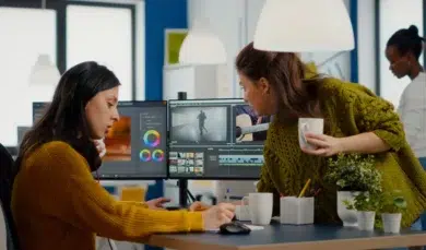Two women collaborating in an office, focused on video editing software on a dual monitor setup. One holds a coffee, creating a productive atmosphere.