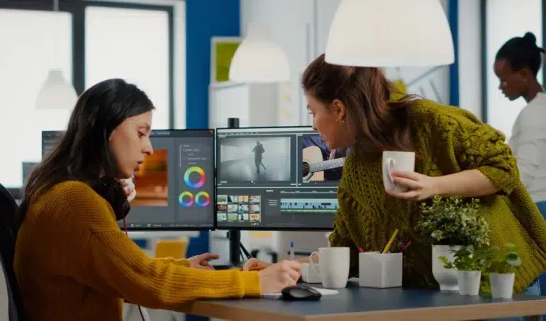 Two women collaborating in an office, focused on video editing software on a dual monitor setup. One holds a coffee, creating a productive atmosphere.