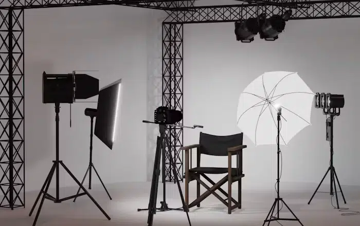 Photography studio setup with lighting equipment, a camera on a tripod, a director's chair, and a white umbrella light against a plain backdrop.
