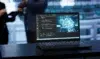 Laptop on a dark table displaying code on the left and a digital brain graphic on the right, with blurred figures in the background.