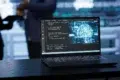 Laptop on a dark table displaying code on the left and a digital brain graphic on the right, with blurred figures in the background.