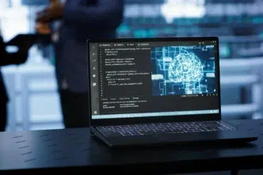 Laptop on a dark table displaying code on the left and a digital brain graphic on the right, with blurred figures in the background.