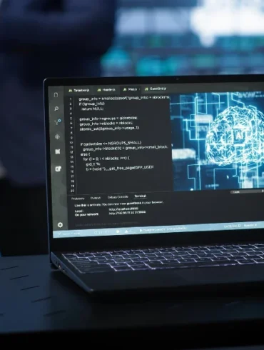 Laptop on a dark table displaying code on the left and a digital brain graphic on the right, with blurred figures in the background.