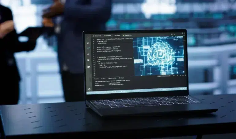 Laptop on a dark table displaying code on the left and a digital brain graphic on the right, with blurred figures in the background.