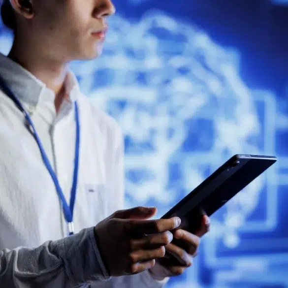 A person in a white shirt holding a tablet, standing in front of a screen displaying a digital brain graphic. The setting is modern and tech-focused.