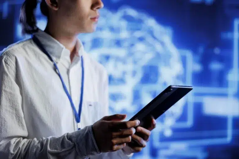 A person in a white shirt holding a tablet, standing in front of a screen displaying a digital brain graphic. The setting is modern and tech-focused.