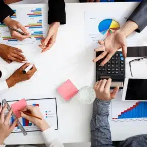 Overhead view of a business meeting with charts, graphs, and a calculator. Hands are gesturing, writing, and using a tablet; a collaborative atmosphere is present.