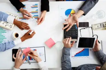 Overhead view of a business meeting with charts, graphs, and a calculator. Hands are gesturing, writing, and using a tablet; a collaborative atmosphere is present.