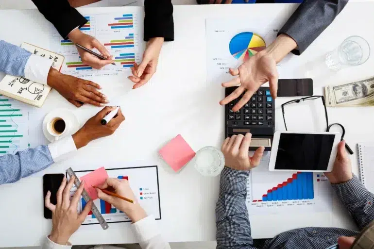 Overhead view of a business meeting with charts, graphs, and a calculator. Hands are gesturing, writing, and using a tablet; a collaborative atmosphere is present.