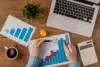 Hands analyze a bar chart with a red arrow, on a wooden desk with a laptop, calculator, coffee, plant, crumpled paper, and another chart. Business context.