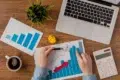 Hands analyze a bar chart with a red arrow, on a wooden desk with a laptop, calculator, coffee, plant, crumpled paper, and another chart. Business context.