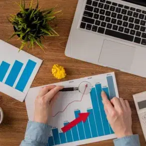 Hands analyze a bar chart with a red arrow, on a wooden desk with a laptop, calculator, coffee, plant, crumpled paper, and another chart. Business context.