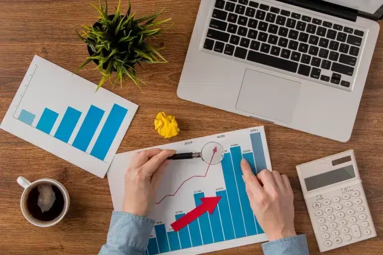 Hands analyze a bar chart with a red arrow, on a wooden desk with a laptop, calculator, coffee, plant, crumpled paper, and another chart. Business context.