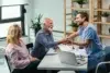 A doctor in scrubs warmly shakes hands with an elderly man in an office setting, while an elderly woman smiles beside him. The mood is friendly and positive.