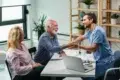 A doctor in scrubs warmly shakes hands with an elderly man in an office setting, while an elderly woman smiles beside him. The mood is friendly and positive.