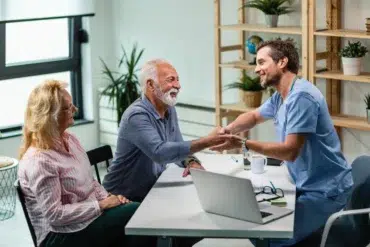 A doctor in scrubs warmly shakes hands with an elderly man in an office setting, while an elderly woman smiles beside him. The mood is friendly and positive.