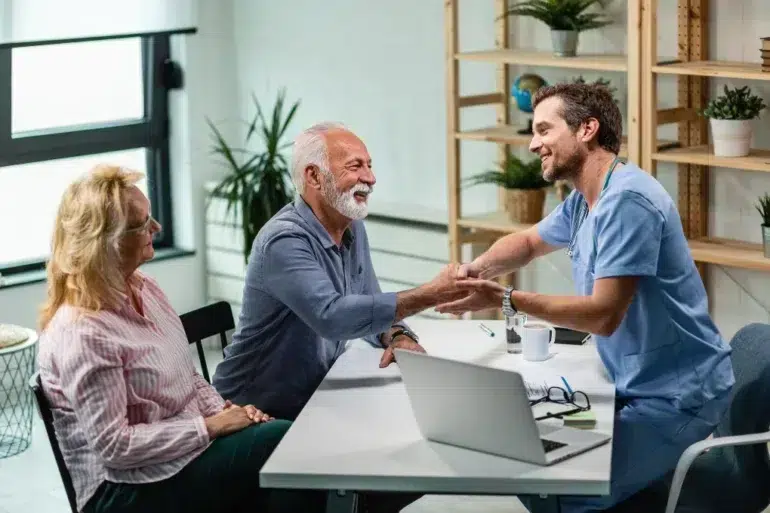 A doctor in scrubs warmly shakes hands with an elderly man in an office setting, while an elderly woman smiles beside him. The mood is friendly and positive.