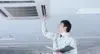 A technician in a light-colored uniform inspects a ceiling-mounted air conditioning unit in an office, holding a clipboard and tool, looking focused.
