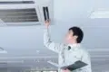 A technician in a light-colored uniform inspects a ceiling-mounted air conditioning unit in an office, holding a clipboard and tool, looking focused.