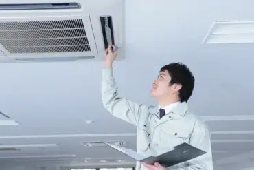 A technician in a light-colored uniform inspects a ceiling-mounted air conditioning unit in an office, holding a clipboard and tool, looking focused.