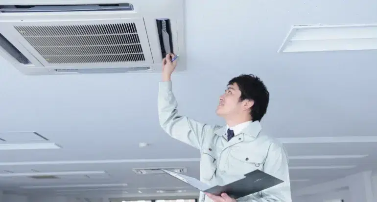 A technician in a light-colored uniform inspects a ceiling-mounted air conditioning unit in an office, holding a clipboard and tool, looking focused.
