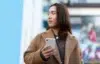 Person in a brown coat holding a white smartphone, standing outdoors with a blurred colorful background.