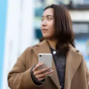 Person in a brown coat holding a white smartphone, standing outdoors with a blurred colorful background.