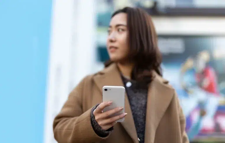Person in a brown coat holding a white smartphone, standing outdoors with a blurred colorful background.