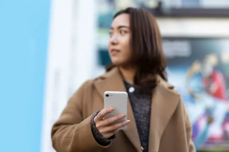 Person in a brown coat holding a white smartphone, standing outdoors with a blurred colorful background.
