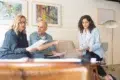 Three professionals discuss documents in a modern office setting with artwork on the wall.
