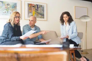 Three professionals discuss documents in a modern office setting with artwork on the wall.