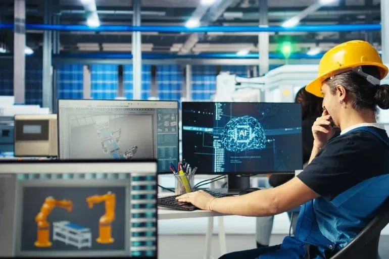 A person in a hard hat and overalls thoughtfully observes computer screens displaying robotic arm designs in a modern industrial setting.