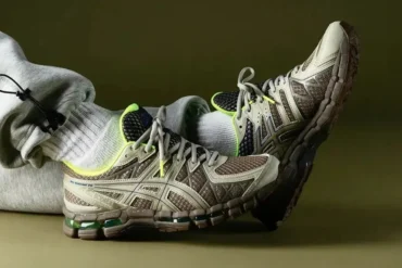 Person wearing gray sweatpants and neon-accented beige athletic shoes, resting feet against a plain olive background.
