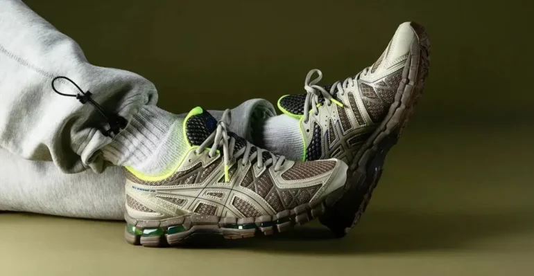 Person wearing gray sweatpants and neon-accented beige athletic shoes, resting feet against a plain olive background.