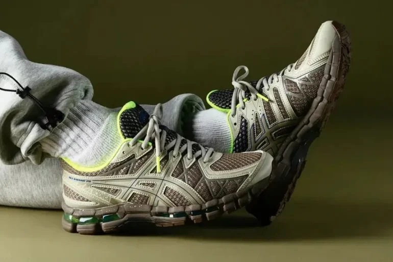 Person wearing gray sweatpants and neon-accented beige athletic shoes, resting feet against a plain olive background.