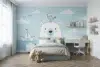 Children's room with a light blue wall featuring a cute bear mural, a bird on its head, and clouds. A cozy bed, desk, and teddy bear add warmth.